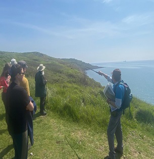 A man gestures out to sea to a group of people standing on a grassy clifftop